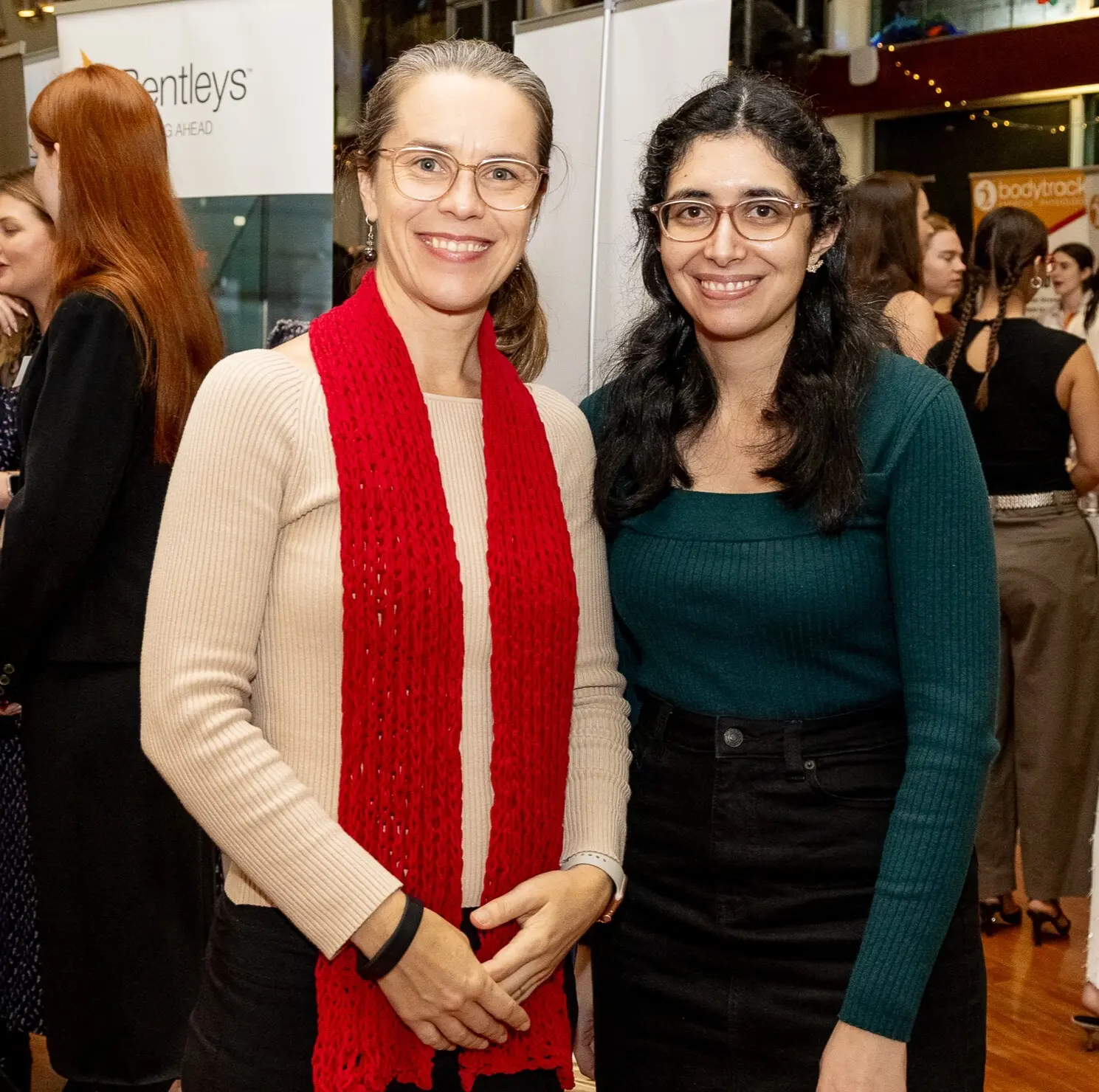 Two women looking at the camera standing in front of a crowded room.